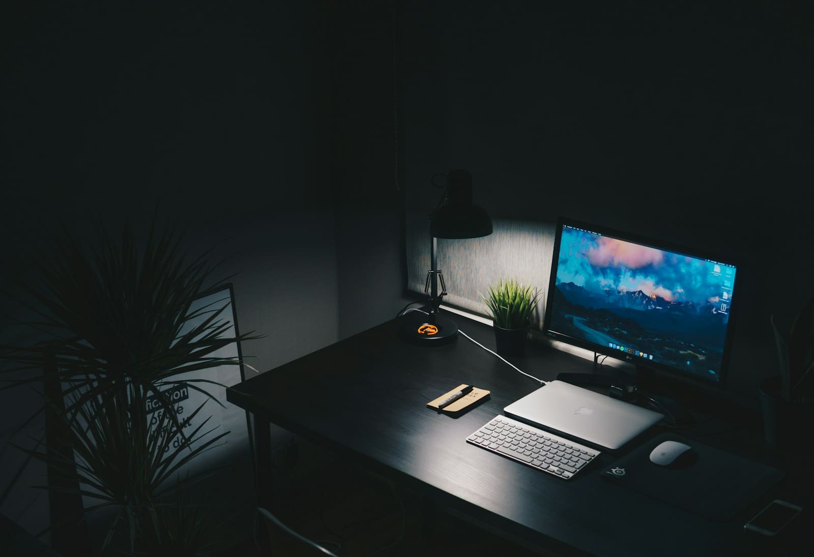 Schreibtisch mit Monitor, Laptop und Pflanzen in gedämpftem Licht